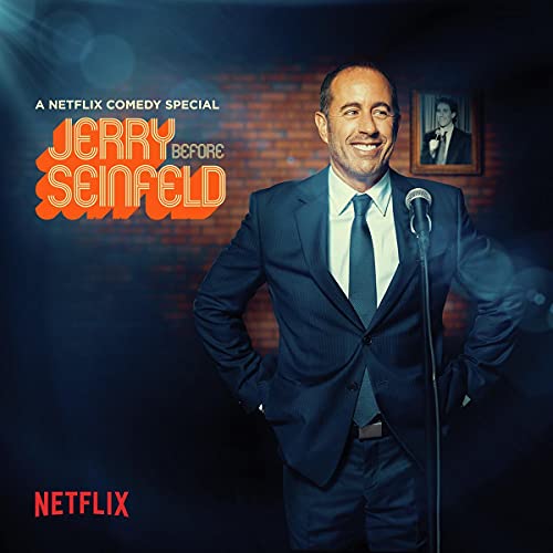 Jerry Seinfeld in a suit at a microphone, with a brick wall backdrop.