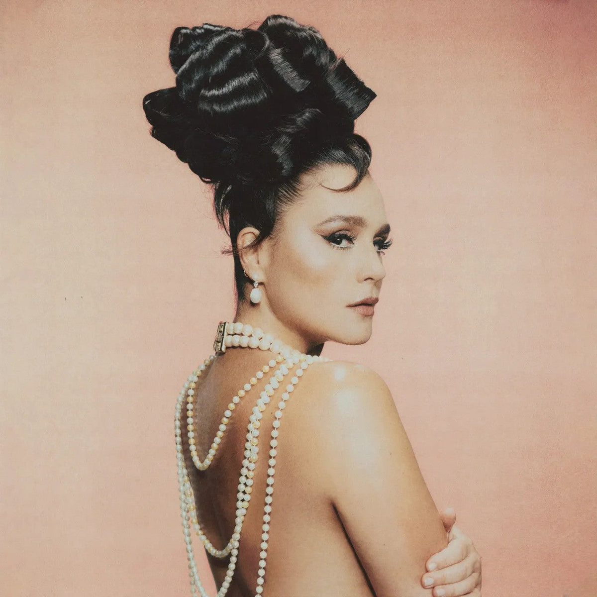 Jessie Ware with an elaborate dark updo and pearl necklaces against a peach background.
