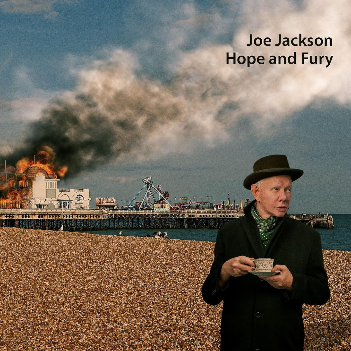 Joe Jackson holding a teacup on a beach with a burning pier behind him.