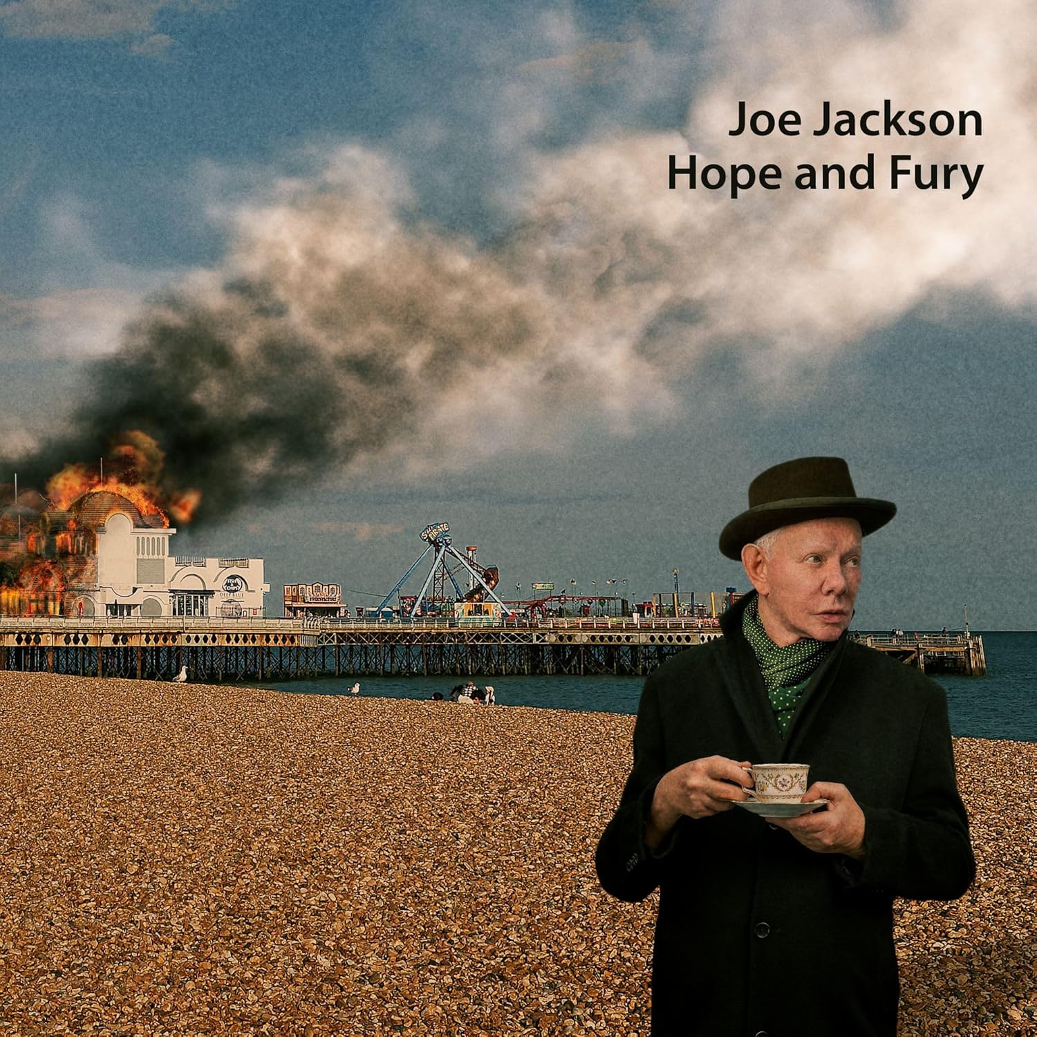 Joe Jackson holding a teacup on a beach with a burning pier behind him.