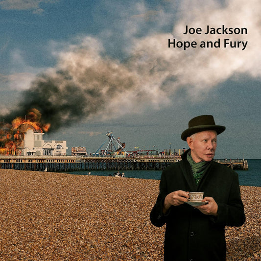 Joe Jackson holding a teacup on a beach with a burning pier behind him.