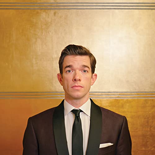 John Mulaney in a suit, centered against a gold textured background.