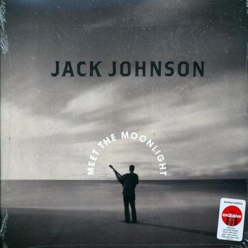 Jack Johnson standing on a beach holding a guitar under a cloudy sky.