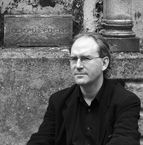 Black and white portrait of a man with glasses, seated near stone ruins.
