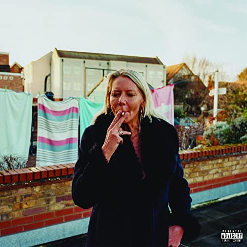 A blonde woman in a black coat smokes a cigarette outdoors near laundry lines.