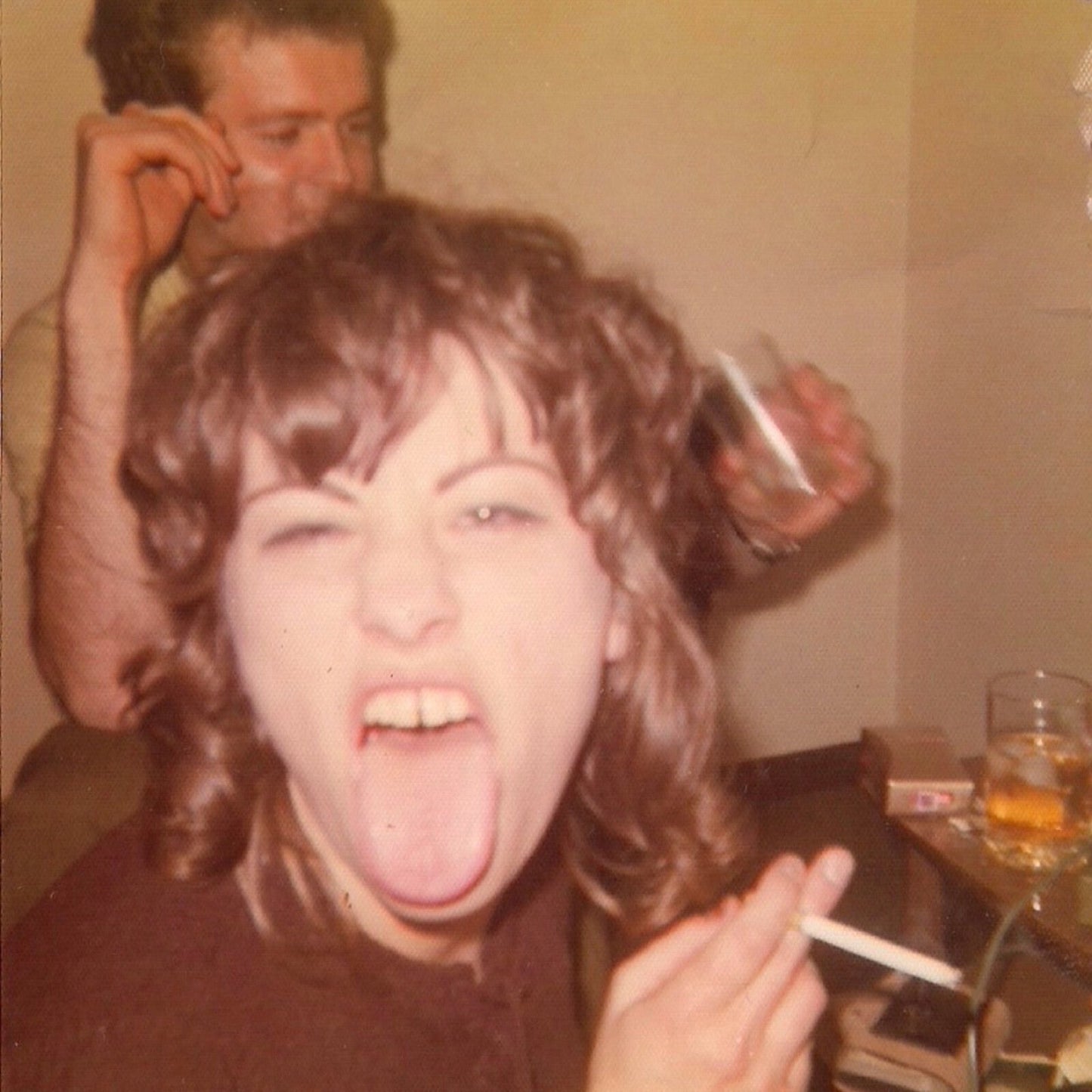 A woman with wavy brown hair sticks her tongue out while holding a cigarette.