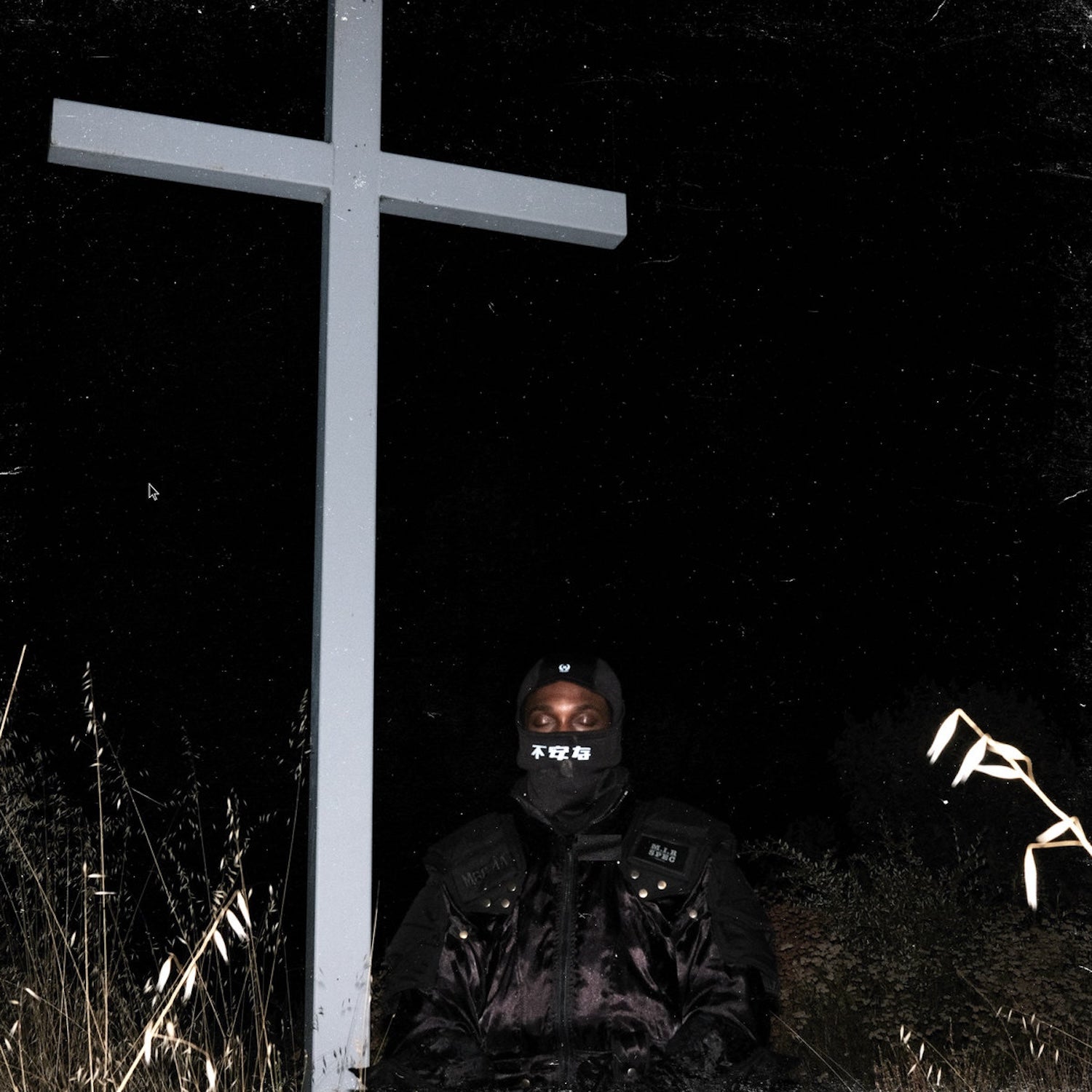 Figure in balaclava kneels before a large, stark white cross in darkness.