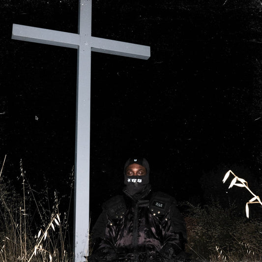Figure in balaclava kneels before a large, stark white cross in darkness.