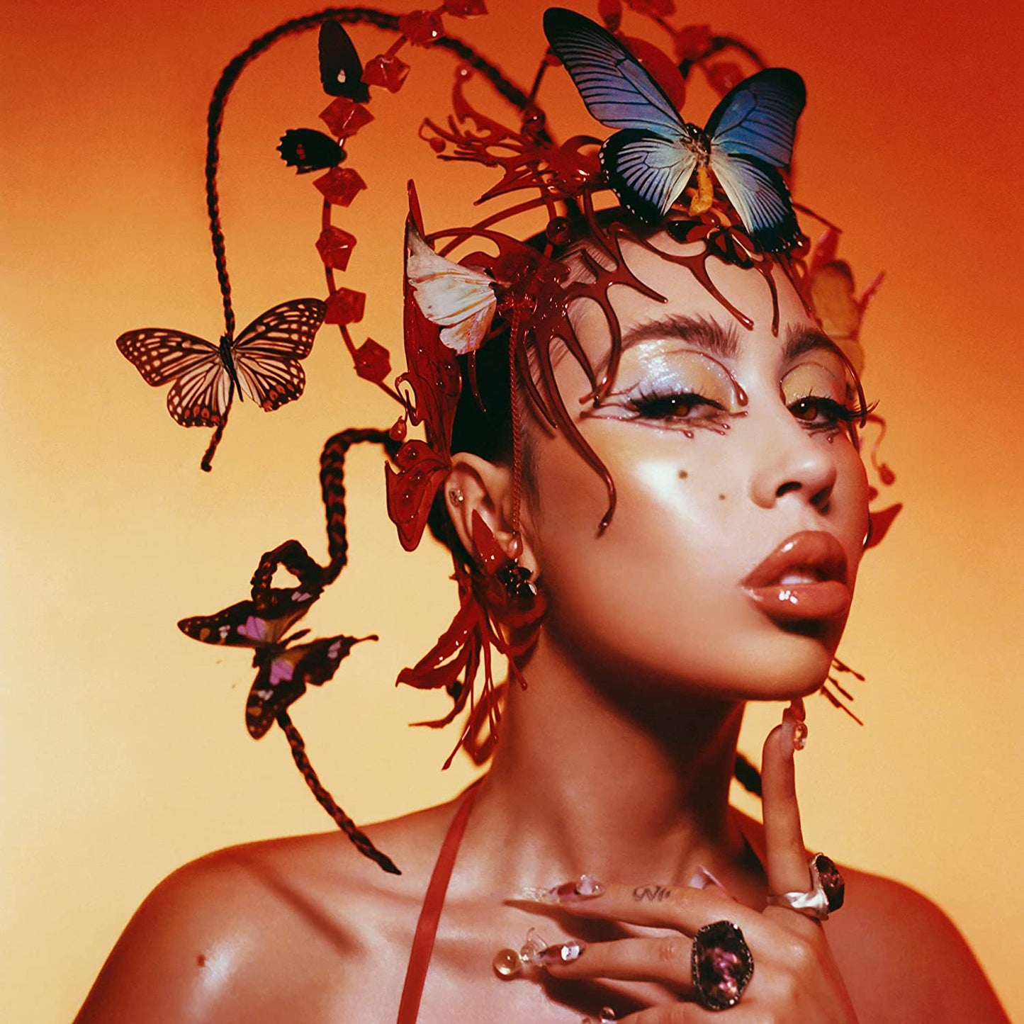 Kali Uchis portrait with butterflies and floral adornments, orange background.