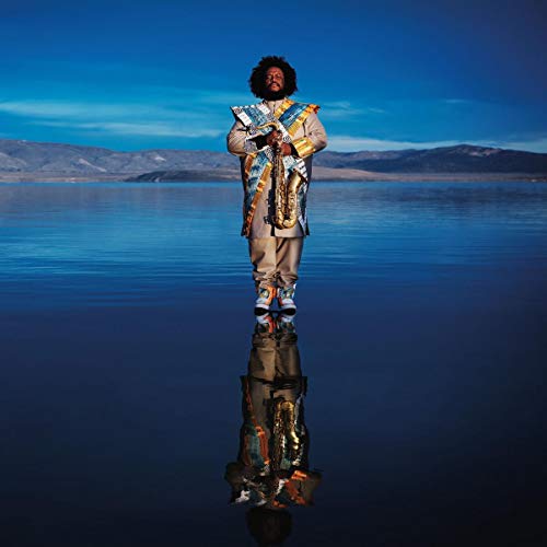 Kamasi Washington stands with saxophone reflected in water, mountains backdrop.