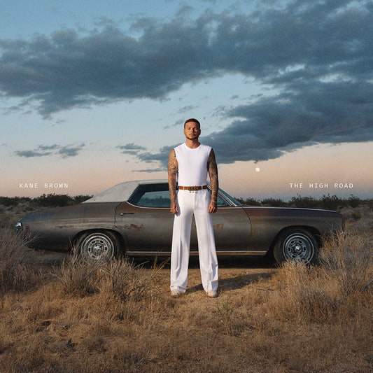 Kane Brown poses beside a vintage car in a dry, grassy field.