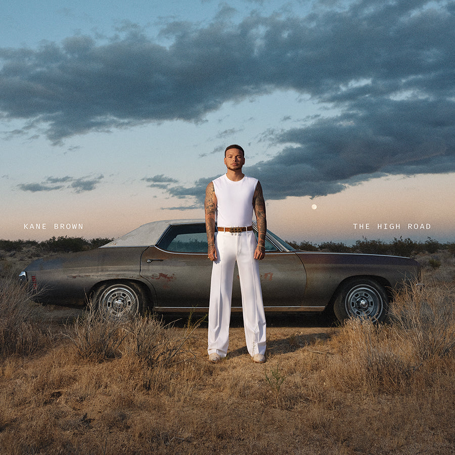 Kane Brown stands beside a vintage car in a dry, grassy field.