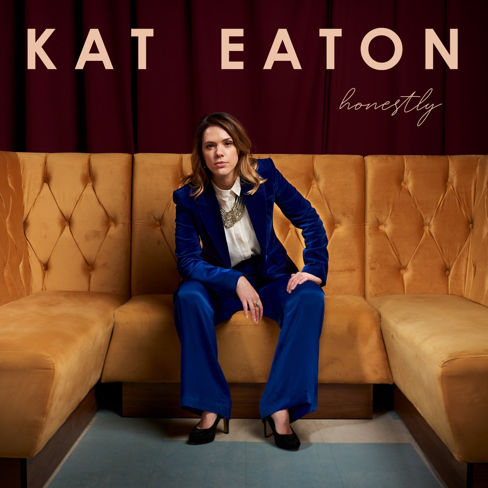 Kat Eaton sits on a gold tufted sofa wearing a blue velvet suit.
