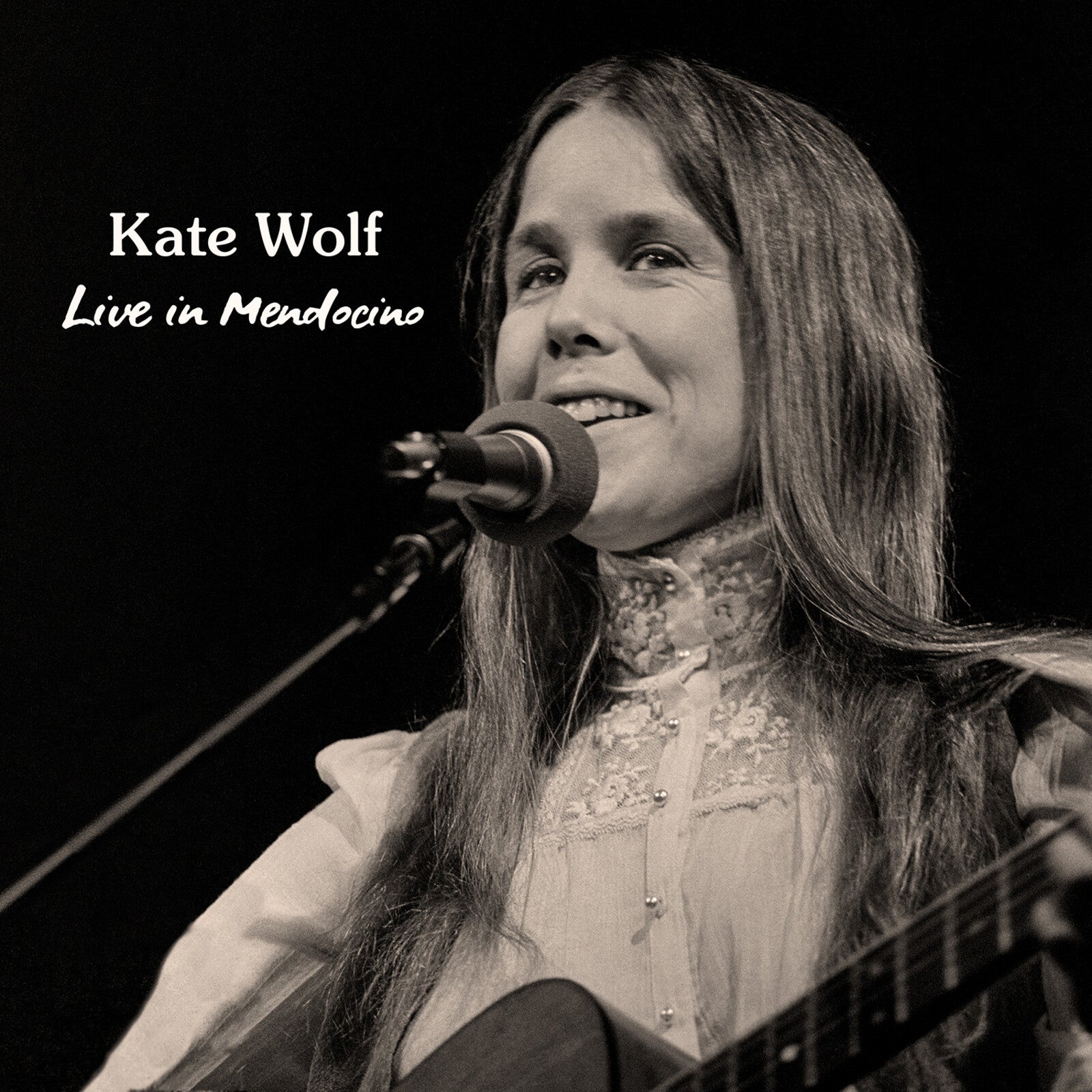 Black and white photo of Kate Wolf singing into a microphone while playing guitar.