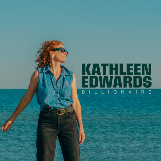 Kathleen Edwards wearing a denim shirt and sunglasses standing before the ocean.