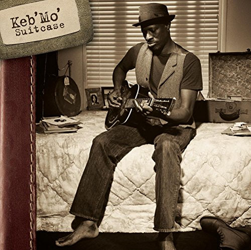 Keb' Mo' sitting on a bed playing guitar in a sepia-toned room.
