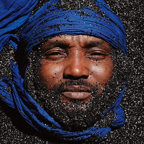 Man with closed eyes, wearing a blue headscarf, buried in dark sand or small rocks.