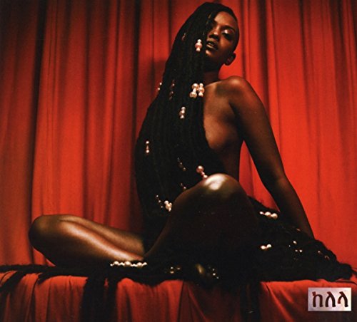 A woman with long braided hair adorned with pearls sits against a red curtain.