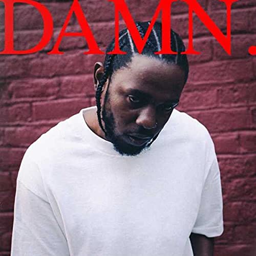 Kendrick Lamar with braided hair wearing a white t-shirt against a red brick wall.