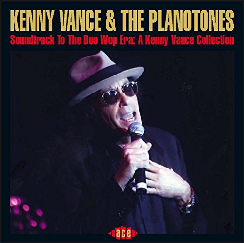 Kenny Vance wearing a hat and sunglasses, singing into a microphone on stage.