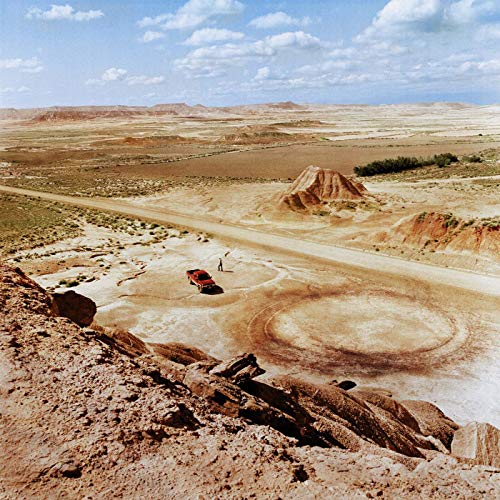 Desert landscape with a red car and circular tire tracks.
