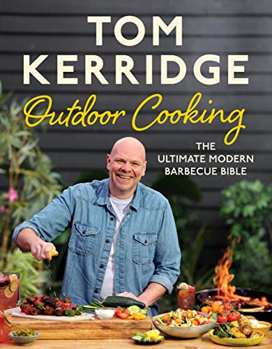 Tom Kerridge smiling while cooking outdoors with various grilled food dishes on a table.