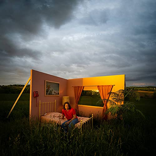 Woman sitting on bed in pink room in grassy field under cloudy sky.