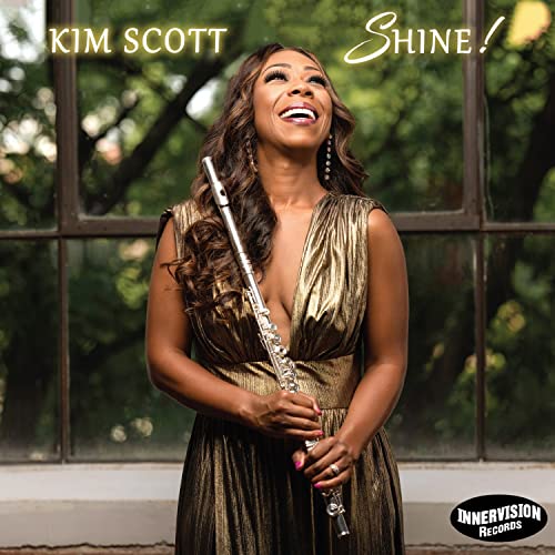 Kim Scott smiling, holding a flute, with a window and trees behind her.