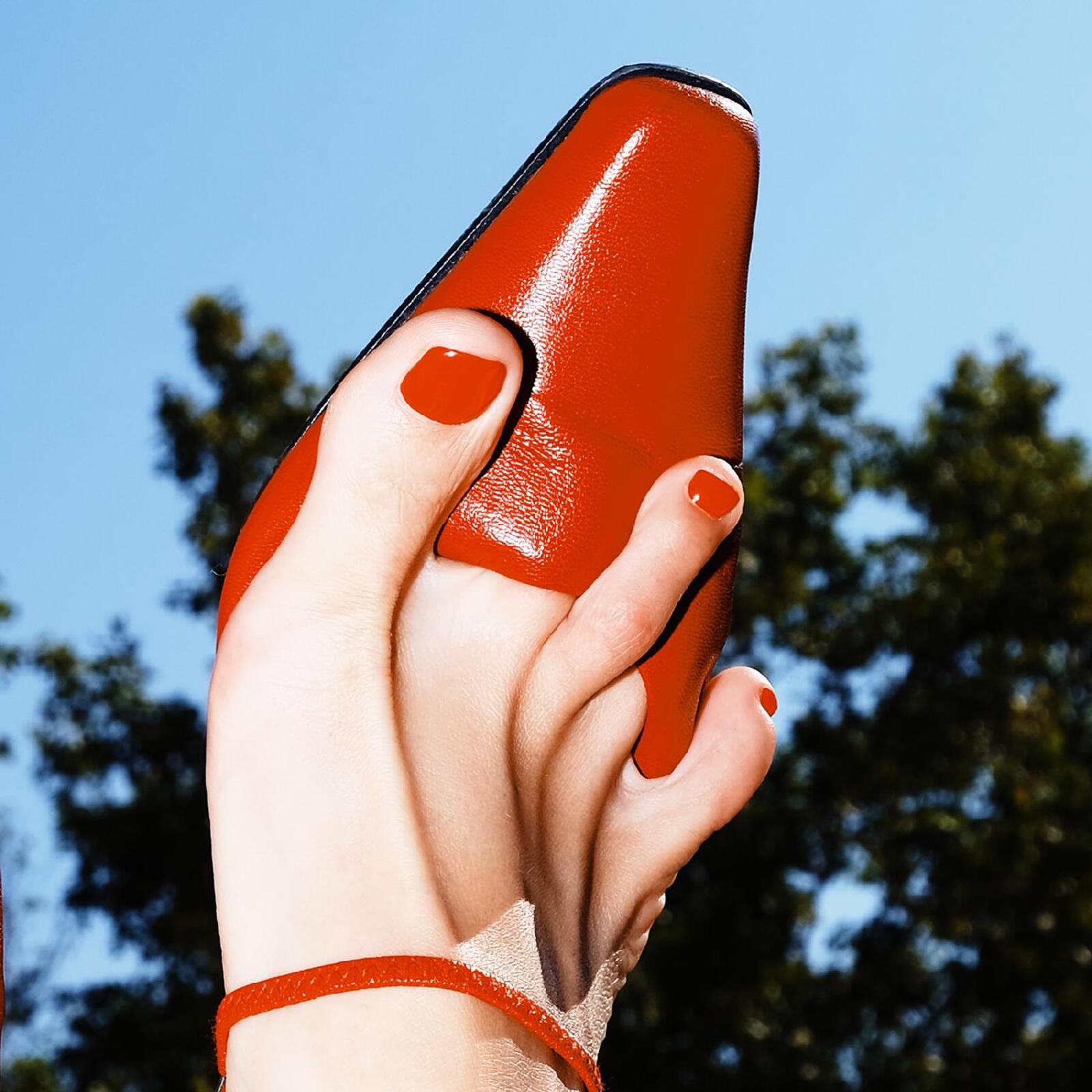 A foot with red nail polish holding a red leather shoe against a sky.