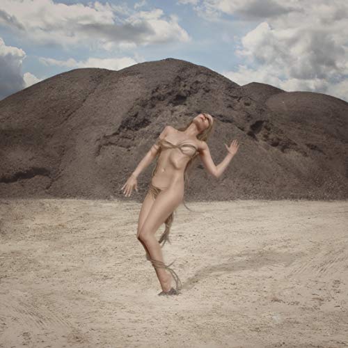 Woman with arms raised stands in a sandy landscape before a dark hill.