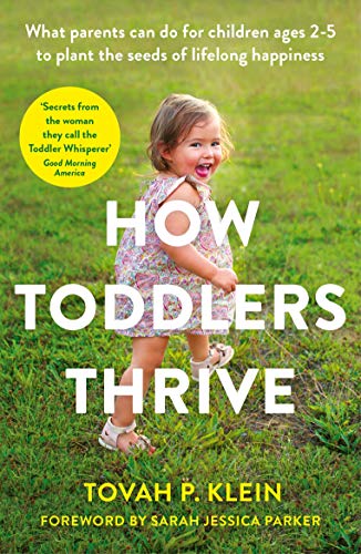 A smiling toddler in a floral dress walks through a grassy field.