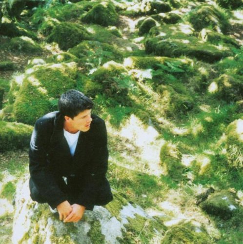 Man in a black coat sits amongst lush green mossy terrain.