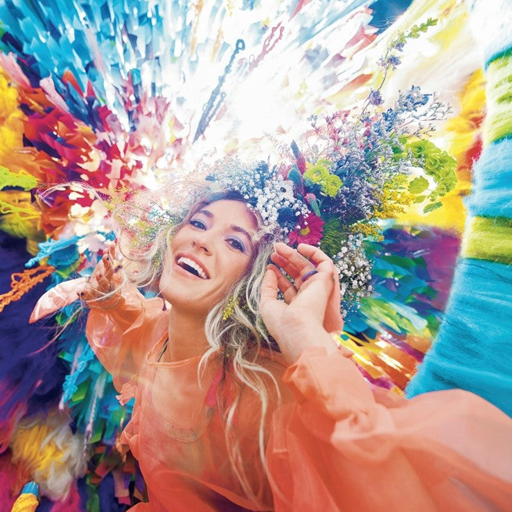 Woman with floral crown, orange dress, surrounded by colorful abstract background, smiling and looking upwards.