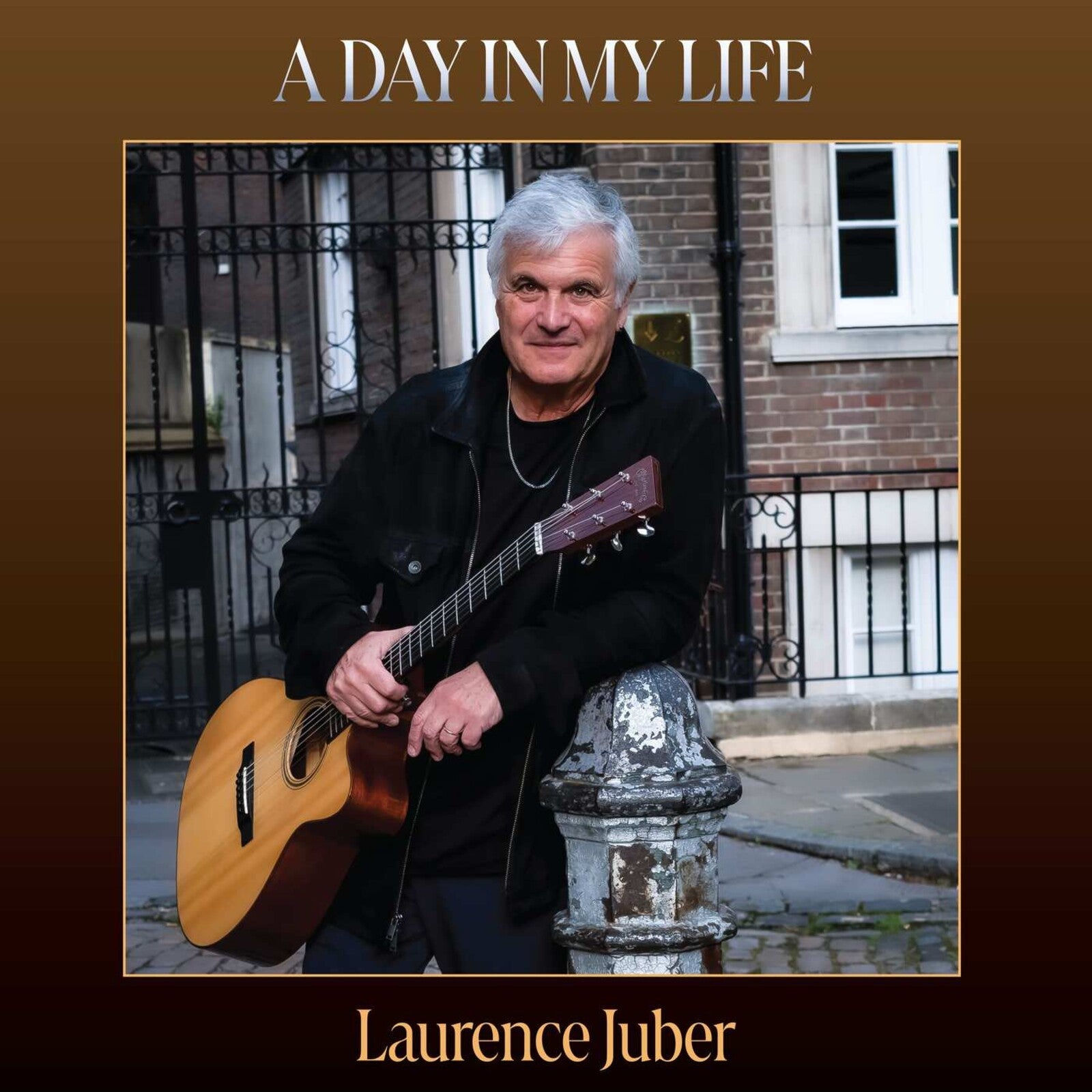 Laurence Juber holding an acoustic guitar, leaning against a stone post on a street.