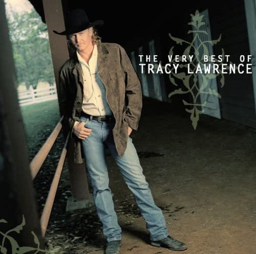 Tracy Lawrence wearing a cowboy hat and jacket standing outside a wooden barn.