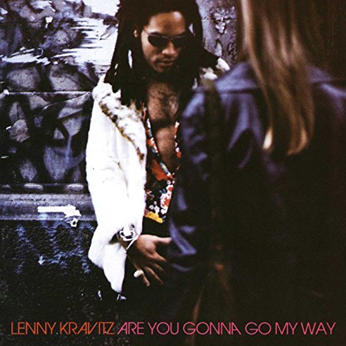 Lenny Kravitz wearing sunglasses and a white fur coat, standing against a graffiti wall.