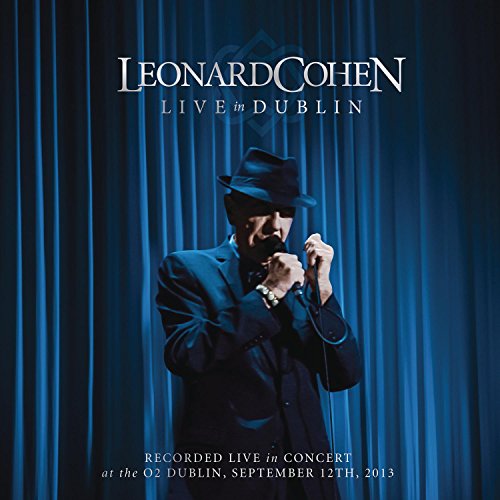 Leonard Cohen in a suit and hat singing into a microphone onstage.