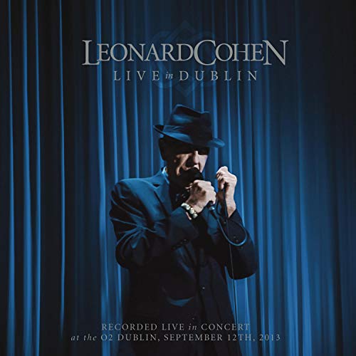 Leonard Cohen wearing a fedora and suit, singing into a microphone before blue curtains.
