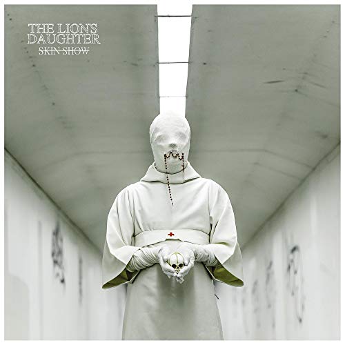 A person in a white mask and robe holds a skull in a tunnel.