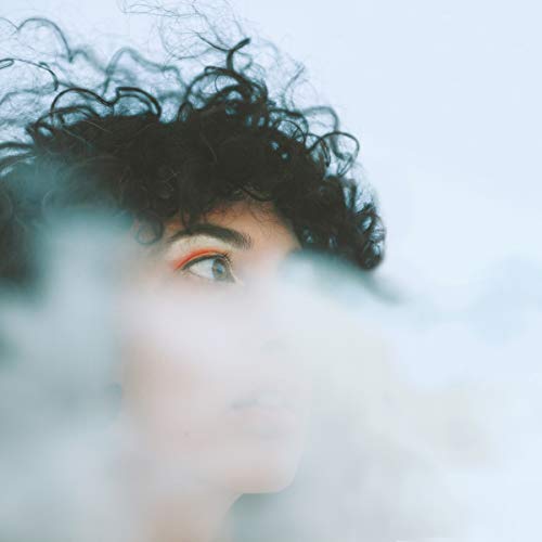A woman with curly black hair and orange eyeshadow, partially obscured by a white, blurry foreground.
