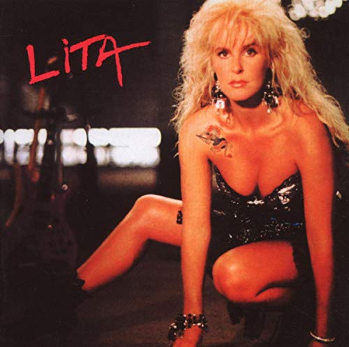 Lita Ford with blonde hair and a tattoo, crouching in a dark, moody setting.