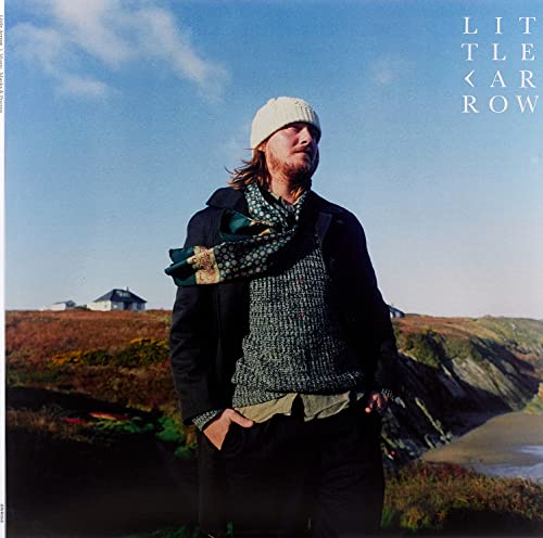 Man in a beanie and coat stands on a coastal hillside.