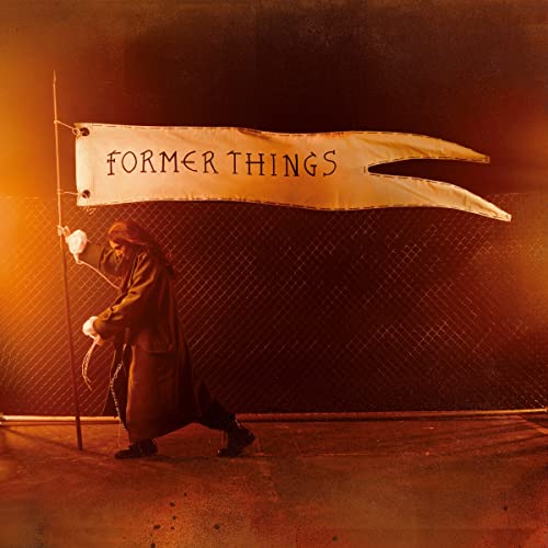 A person in a dark coat holds a banner reading Former Things against chain-link fencing.