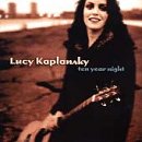 Lucy Kaplansky album cover: woman with guitar on rocky shore with cityscape.