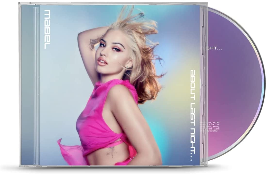 Blonde woman in pink outfit with one arm raised, hair blowing, against colorful background.