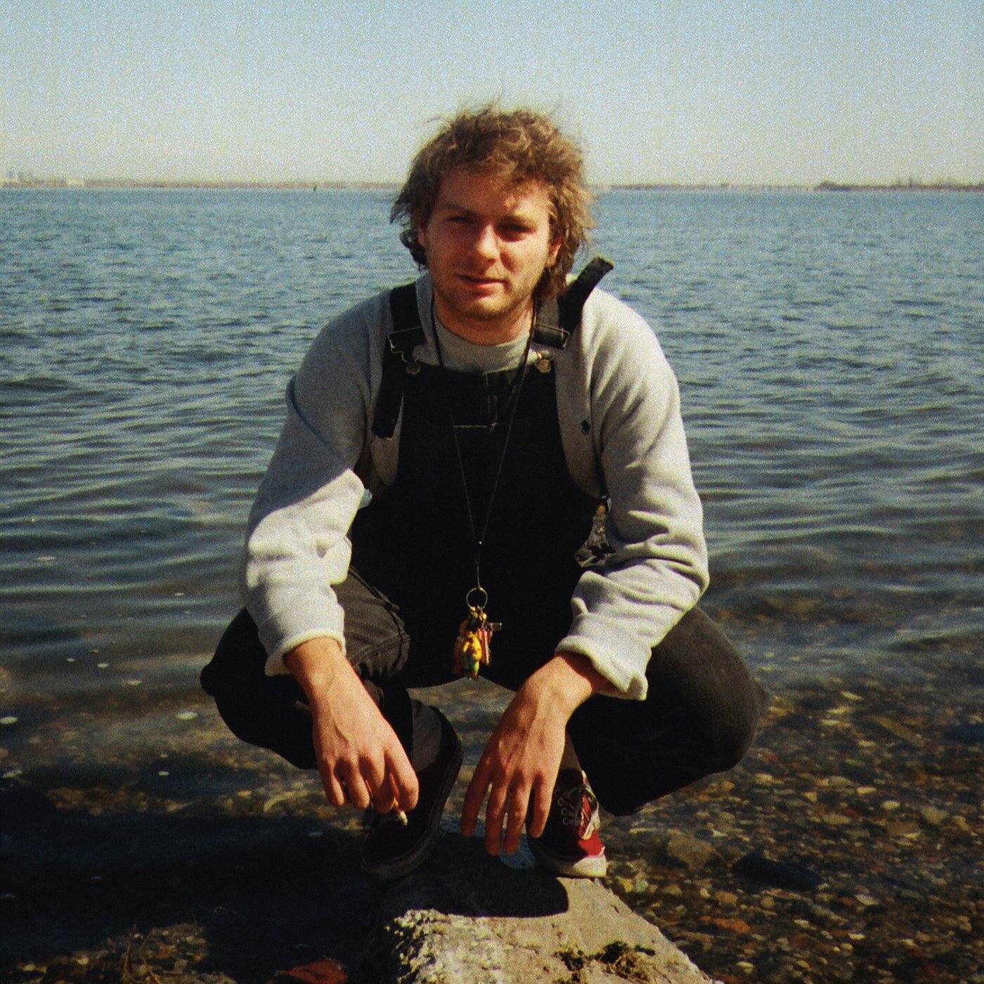 Mac DeMarco squats by water, wearing overalls and a grey sweater.