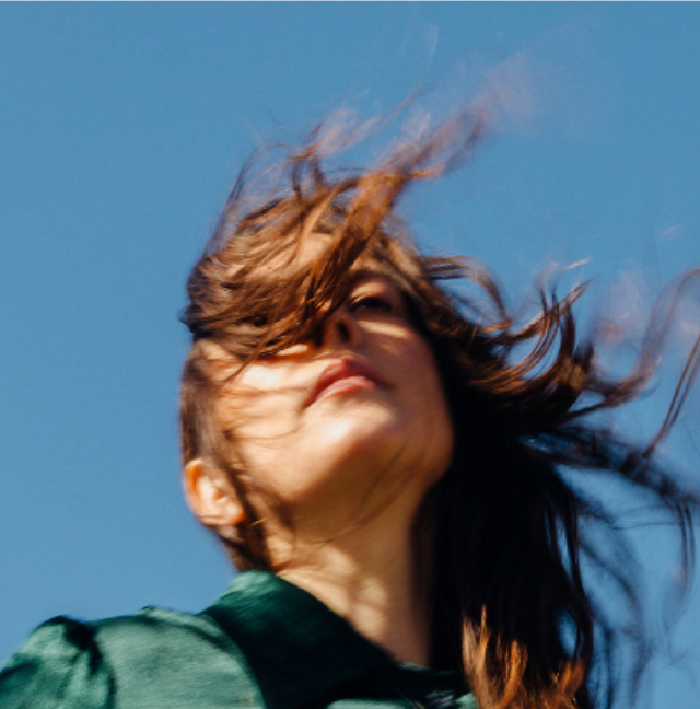 A woman with windblown hair looks upward against a clear blue sky.