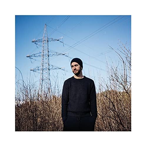 Man in black sweater stands before power lines and dry grass.