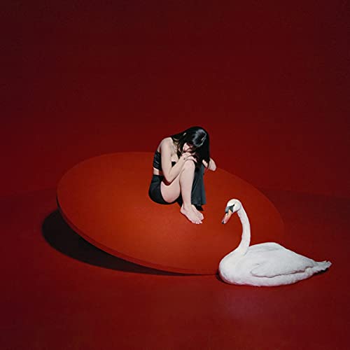 Woman with knees drawn up sits beside a white swan on red.
