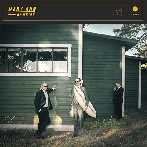 Three men in suits and sunglasses stand before a green wooden building.